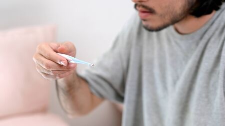 Closeup of man looking at thermometer, Male hand holding a digital thermometer when measures the temperature, People get fever, flu from virus check body temperature when stay at home, Sick people and medical instrumentの写真素材