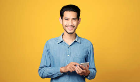 Asian man holding mobile phone smiling while standing isolated on yellow background, People on...