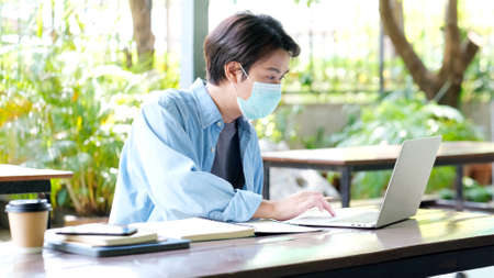 Young asian campus student man wearing protection mask working with laptop computer while online...