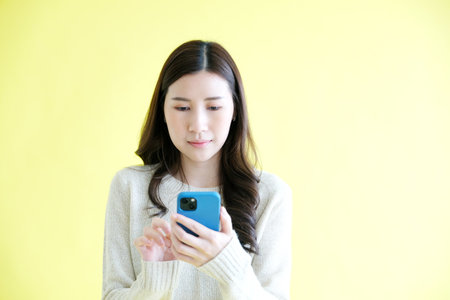 Young woman holding mobile phone while standing over isolated yellow backgroundの写真素材