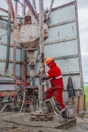 Gydan Peninsula / Russia - August 11 2019: Work driller in red uniform, in helmet and goggles. He installs drill pipes for screwing and lowering them into an oil well, and continuing its drilling. The concept of a working person.のeditorial素材