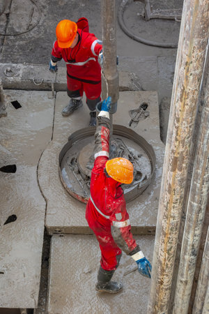 Two working drillers in a red uniform, in a helmet and goggles, install drill pipes after lifting them from an oil well after drilling. The concept of a working person.のeditorial素材