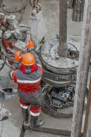 Work driller in red uniform, in helmet and goggles. He uses a hydraulic wrench to screw drill pipes to lower them into an oil well and continue drilling it. The concept of a working person.のeditorial素材