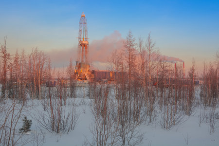 A rig for drilling oil and gas wells in the northern landscape of the forest-tundra. The rising winter sun colors the landscape orange. Puffs of steam and smoke partially cover the oil derrickの写真素材