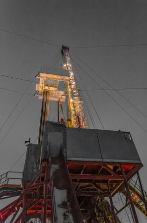 Industrial drilling rig silhouetted against dusky winter sky, with snow-covered ground and camp...