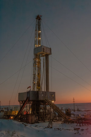 Industrial drilling rig silhouetted against dusky winter sky, with snow-covered ground and camp...