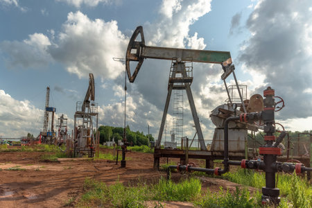Oil production at the field using two rocking machines. In the background is a drilling rig and equipment for drilling wells. Summer. Beautiful sky and natureの写真素材