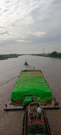 Barge and tugboat on the river, laden with cargo covered in green tarpaulin under cloudy skiesの写真素材