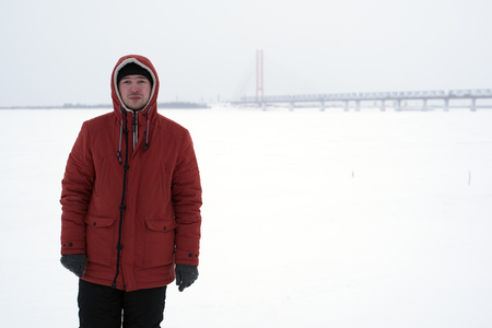 The young man in the orange jacket and black pants, standing next to a frozen river near the bridgeの写真素材