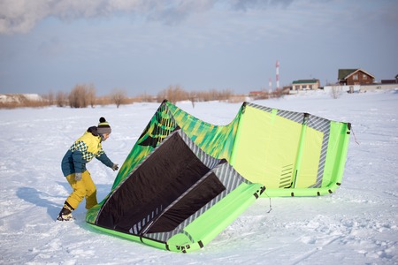 The athlete in the winter is preparing a green kite for Snowkiting on the snowy field