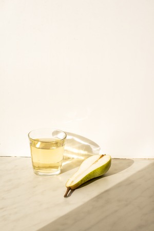 Transparent glass Cup with pear lemonade and pear under natural sunlight and shade on marble surface. The shadow falls on a white wall.の写真素材