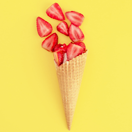 Ice cream with red strawberries divided in half and red sweets in waffle cone on yellow surfaceの写真素材
