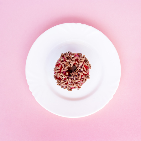 Donut covered with red glaze and sprinkled with grated chocolate on white plate on pink backgroundの写真素材