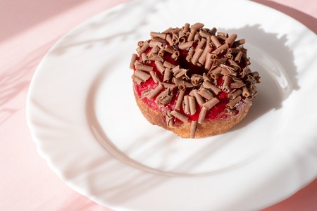 Donut covered with red glaze and sprinkled with grated chocolate on white plate on pink background...
