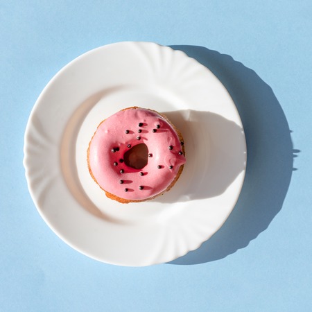 Donut covered with pink frosting and sprinkled with black roundels on a white plate on a blue background under natural sunlightの写真素材