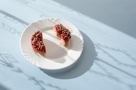 Donut cut in half with red glaze and sprinkled with grated chocolate on white plate on blue background under natural sunlight with patterned shadowsの写真素材