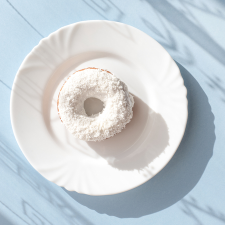 Donut covered with white glaze and sprinkled with coconut flakes on a white plate on a blue background under natural light with shadows of patterns on the surfaceの写真素材