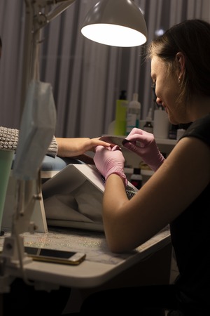 In the nail salon client manicurist in pink gloves handles nails with nail filesの写真素材