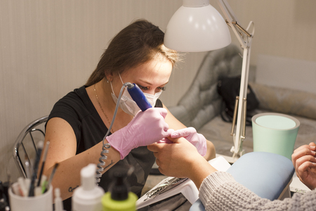 In the nail salon client manicurist in pink gloves removes varnish with blue manicure deviceの写真素材
