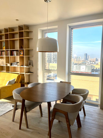 A lifestyle photo of open space dining room with a wooden table, chandelier and chairs. And living room with a yellow couch, chair and a wall-sized bookshelf. European apartment.の写真素材