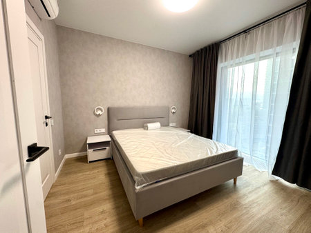 Freshly renovated modern bedroom with a grey bed, new mattress in plastic, and minimalist decor. Features wall lights, a nightstand, and large window with curtains.の写真素材