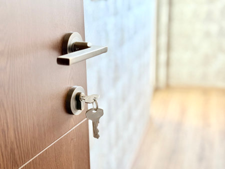 A close-up of a wooden door with a modern metal handle and a key in the lock, slightly ajar. It reveals a softly lit interior with wooden flooring, creating a sense of entryの写真素材