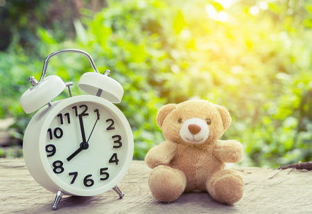white alarm clock and teddy bear on wood table and blurred background  in morning with sun light and vintage filterの写真素材