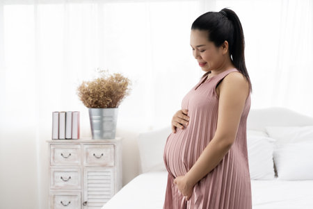 A beautiful Asian pregnant woman standing happily smiling and touching the baby in the womb in the white bedroom.の写真素材