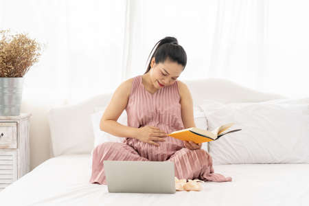 Pregnant women are reading books for their children in the belly in the white bedroom. Pregnant in the third quarter. For the development of the baby.の写真素材