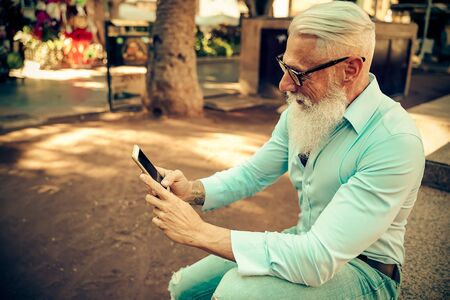 Smiling senior man relaxing in the park. Mature male having fun with new technology trends. - Focus...