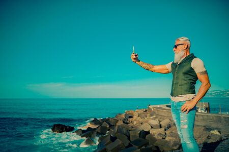 Trendy senior man using smartphone app near the sea - Mature fashion male having fun with new trends technology - Tech and joyful elderly lifestyle concept - Focus on his face - Imageの写真素材