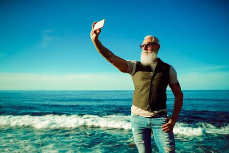 Trendy senior man using smartphone app near the sea - Mature fashion male having fun with new trends technology - Tech and joyful elderly lifestyle concept - Focus on his face - Imageの写真素材