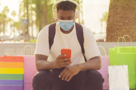 Boy waiting for his girlfriend during shopping. Man listening the music in the commercial center with protective mask for coronavirus. Imageの写真素材