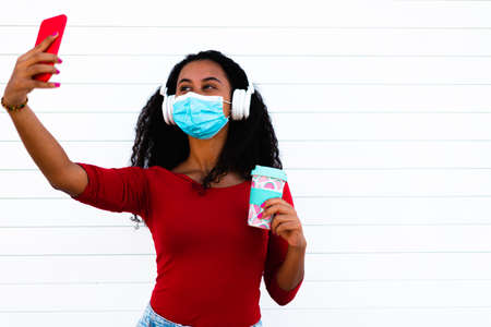 Young latin american woman using mobile phone for selfie and listening music. Girl wearing face mask for coronavirus prevention. Technology and virus concept.の写真素材
