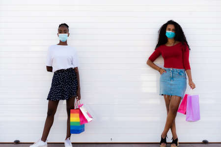 Spring shopping during quarantine conception: fashionable women wearing protective mask posing with colorful paper bags. White background. Copy, empty space for textの写真素材
