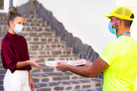 Delivery man employee in yellow cap and t-shirt uniform face mask hold food order pizza box. Service quarantine pandemic  virus flu   concept.の写真素材