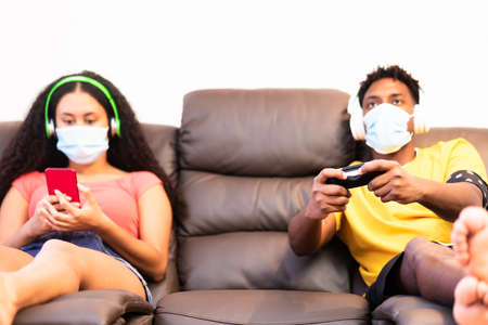 Boy having fun with video games console, girl looking the smartphone. Young millennial people with protective mask  .の写真素材