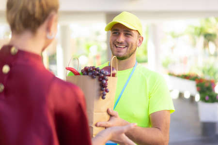 Handsome food delivery service man in yellow shirt holding fresh fruits and vegetables to customer at near home. Express delivery, takeaway food delivery and online shopping concept.の写真素材