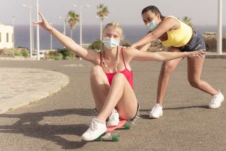 Two female friends with protective masks playing with skateboard. One girl pushing other from behind. Friendship and coronavirus concept.の写真素材