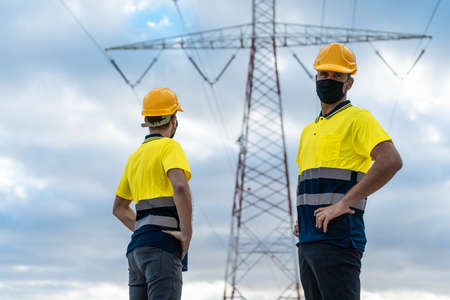 Electrical engineers working. Electrician looking the camera and another skilled worker looking high voltage tower against blue sky.の写真素材
