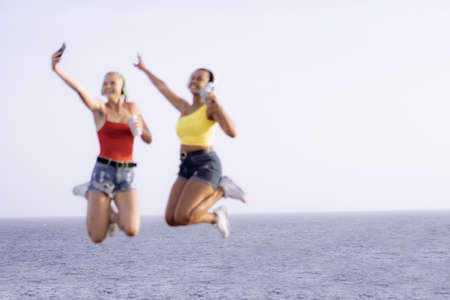 Two happy joyful young women jumping and laughing together over sea background. Defocused people. Copy space.の写真素材