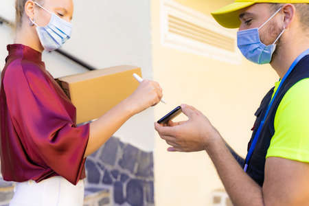 Caucasian woman with protective mask appending signature sign on smart phone after accepting receive box from delivery man, woman sign on the smartphone. Delivery and coronavirus concept.の写真素材