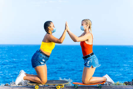 Two female friends with protective masks playing with skateboard. Girlfriends celebrating their success, high fiving Friendship and coronavirus concept.の写真素材