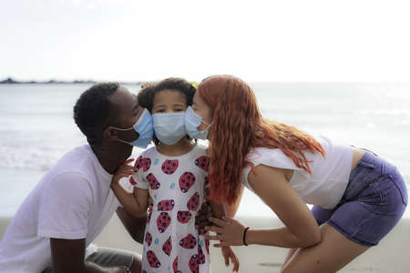 Multiethnic family kissing next to the sea while wearing a face mask that protects them from infection because of the outbreak of virus infection COVID-19.の写真素材