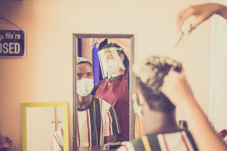 Adult african man at hairdresser wearing protective mask due to coronavirus pandemic. Young asian woman hair cutting in covid time.の写真素材
