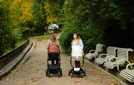 Two happy mothers with their baby strollers walking together in park. Prams for newborns. Motherhood