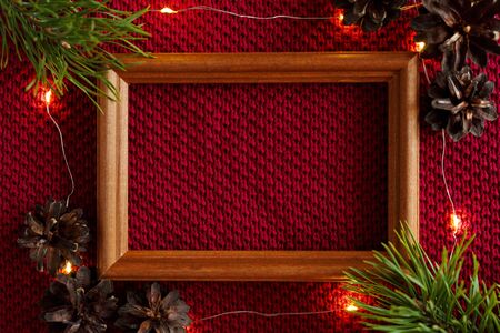 Christmas background. Cones, a wooden frame and a garland lying on a red knitted texture of a woolen...