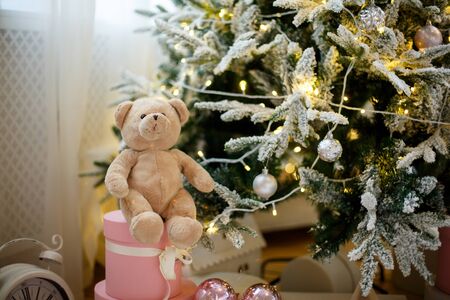 A teddy bear toy is sitting on a pink gift box, at the Christmas tree decorated with yellow garlands and ballsの写真素材