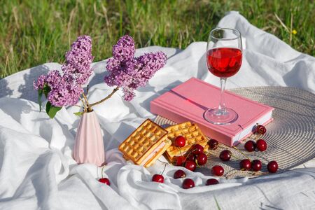 Still life and food photo. Cherry and waffle berries lie on a gray wicker round napkin and white wave-crumpled fabric. A glass of cherry juice or wine. Vase with lilac flowers.の写真素材