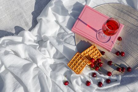 Still life and food photo. Cherry and waffle berries lie on a gray wicker round napkin and white wave-crumpled fabric. A glass of cherry juice or wineの写真素材